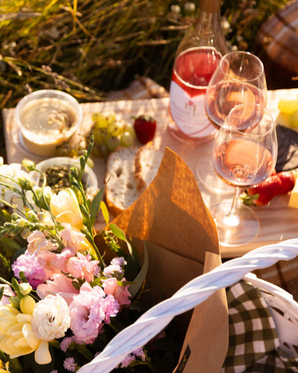 Outdoor picnic setup with wine, cheese, and flowers on a checkered blanket.